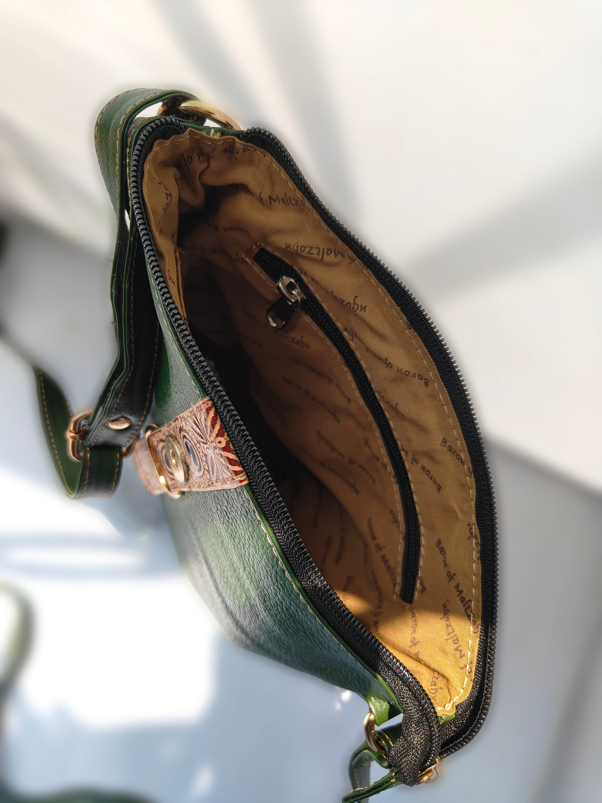 A close-up of a dark green leather shoulder bag shown open, displaying a tan patterned interior lining with a black zipper and a small decorative leather detail with a rose gold-toned emblem.