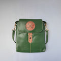 A green leather crossbody bag with a flap closure, featuring a circular orange-brown tooled ornament on the front and a matching vertical tooled accent piece.