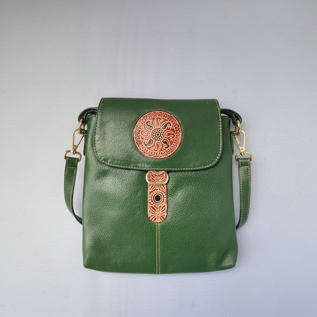 A green leather crossbody bag with a flap closure, featuring a circular orange-brown tooled ornament on the front and a matching vertical tooled accent piece.