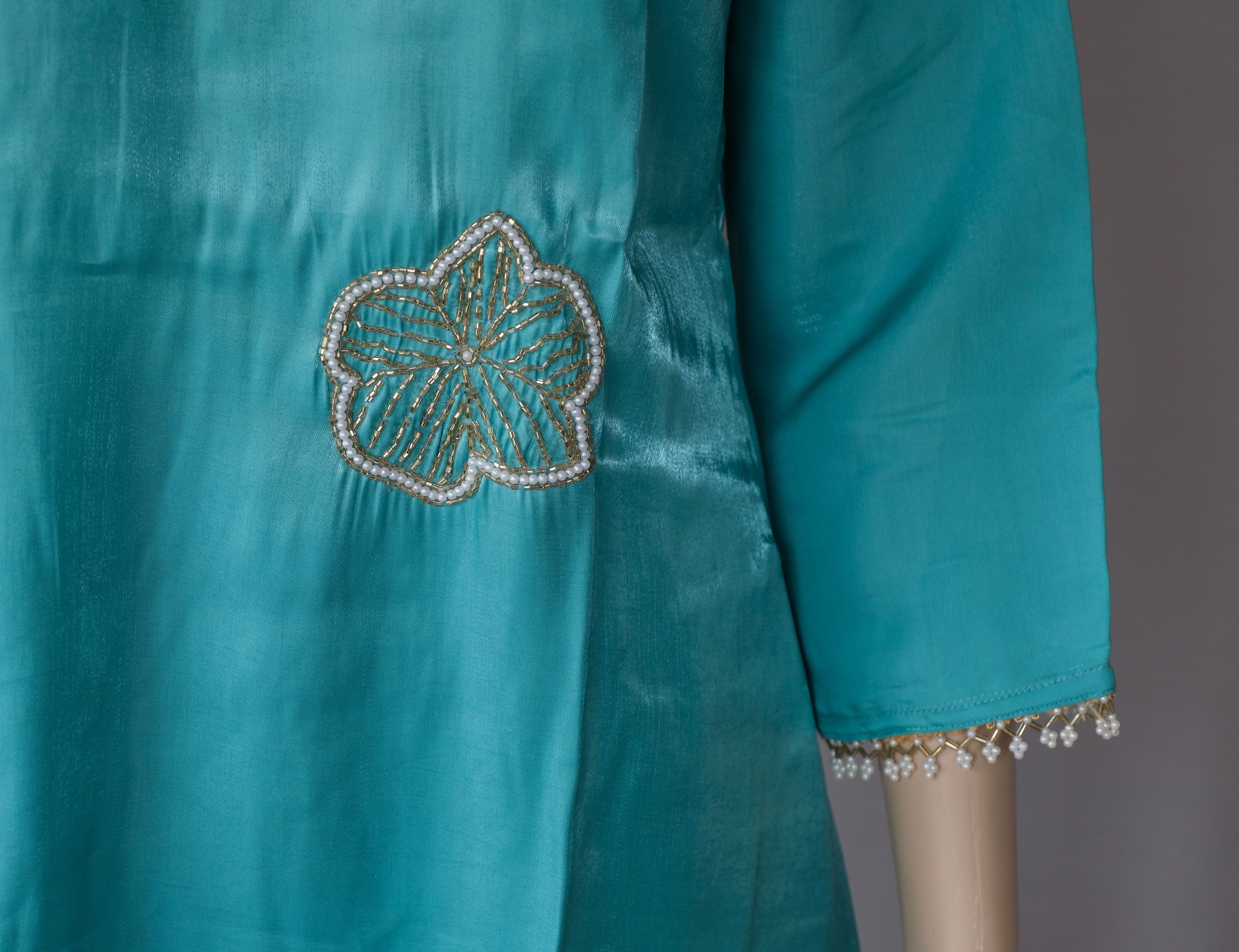 Close-up of a teal kurta featuring a floral embroidered patch with gold and white threadwork and bead trim on the sleeve cuff.
