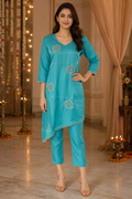 A woman modeling a turquoise blue kurta set with matching pants in an indoor decorated setting with warm string lights and curtains.