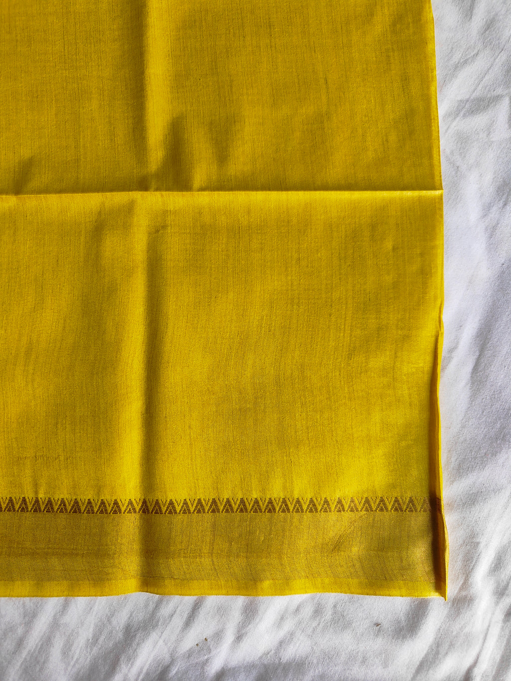 A folded yellow fabric, likely a saree or textile, laid flat on a white wrinkled surface. The fabric features a decorative border of small black triangles along the bottom edge.