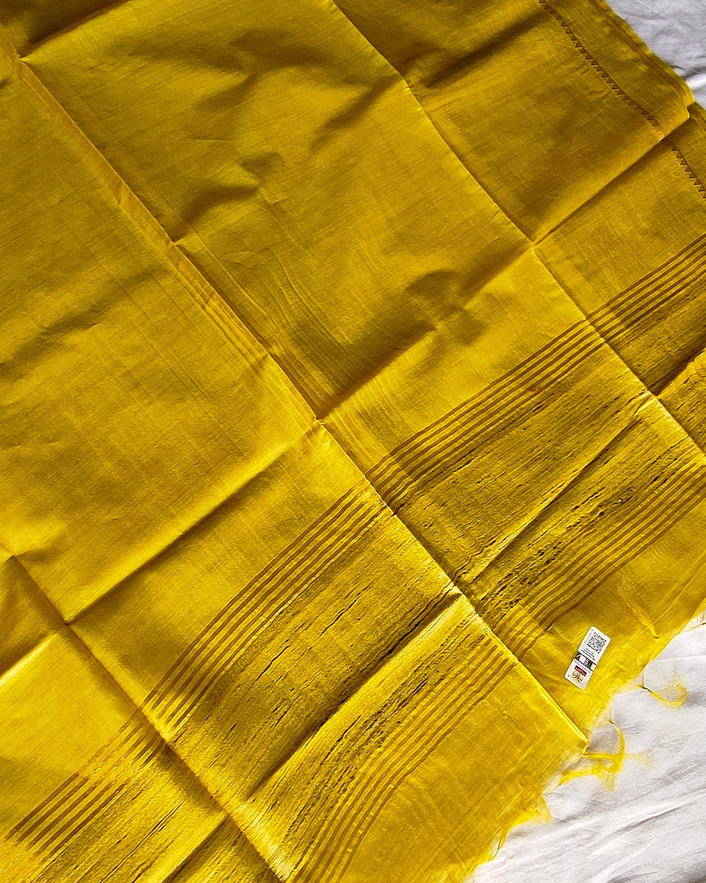 A bright yellow fabric, likely a sari, is laid out flat on a white sheet. The fabric features a subtle woven texture with fine horizontal stripes and a small barcode label near the top right.