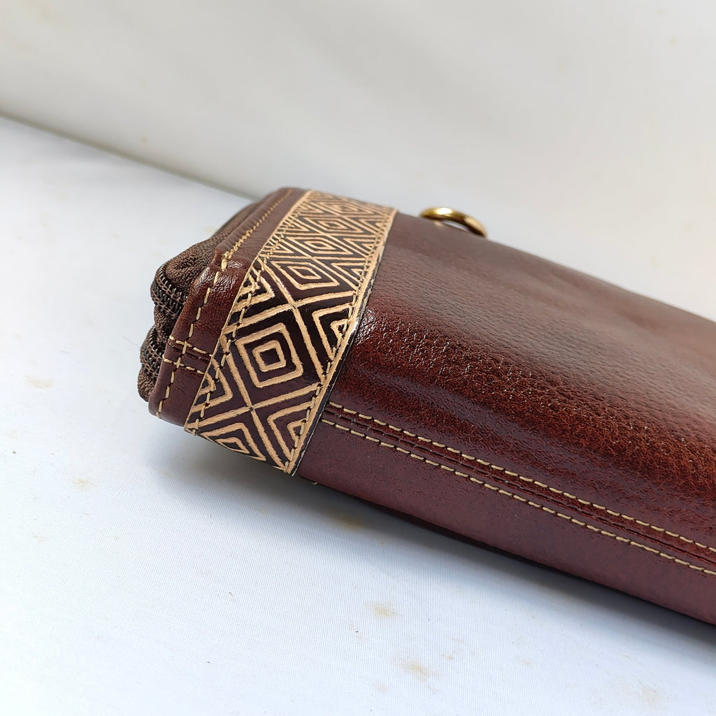 A close-up of a brown leather pouch or wallet showing a decorative geometric patterned band in dark brown and tan, with visible stitching and a small gold-tone ring detail, resting on a white surface.