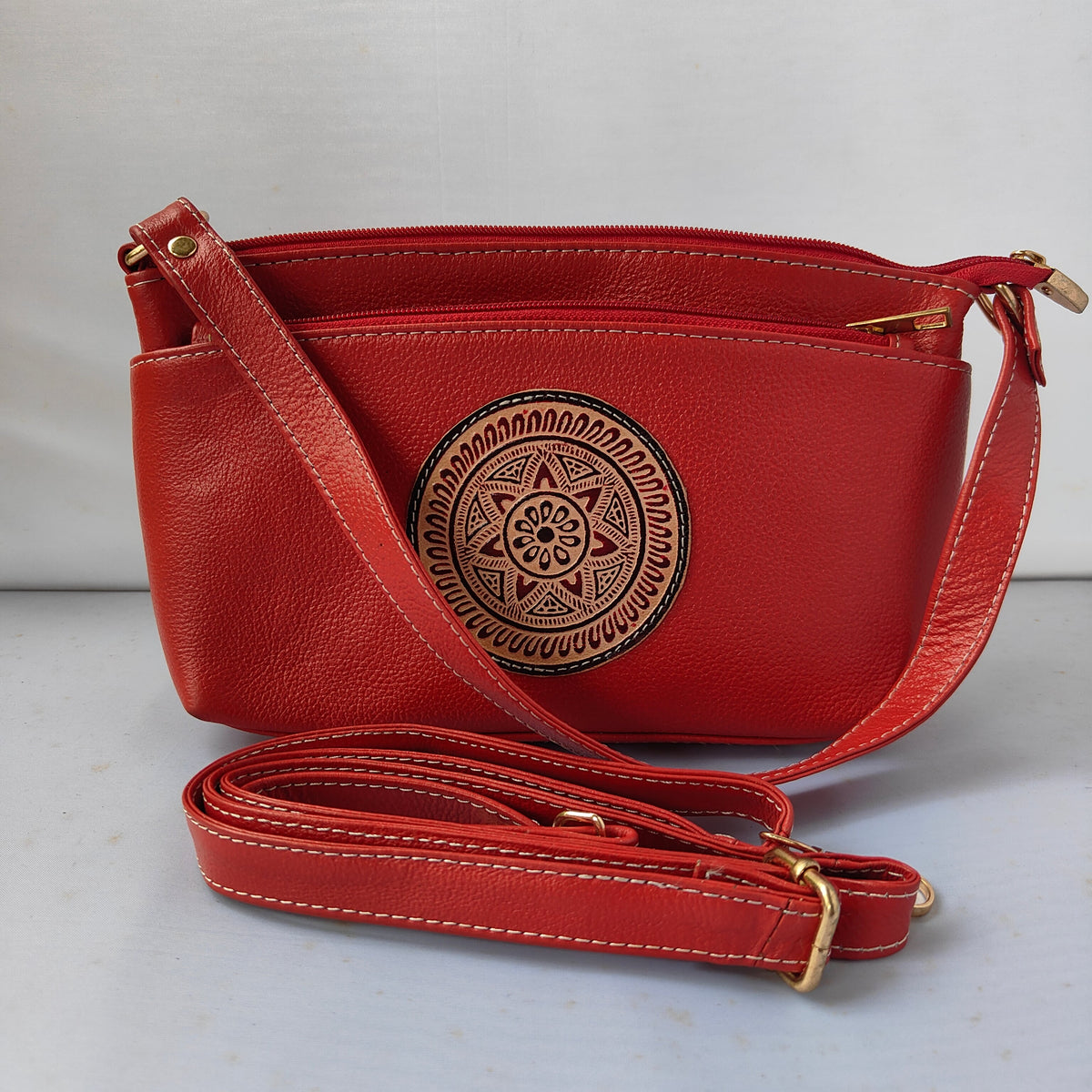 A red faux-leather crossbody handbag with a zippered top, a circular brown embossed tribal-style emblem on the front, and a detachable red shoulder strap with gold-tone hardware, displayed on a light gray surface.