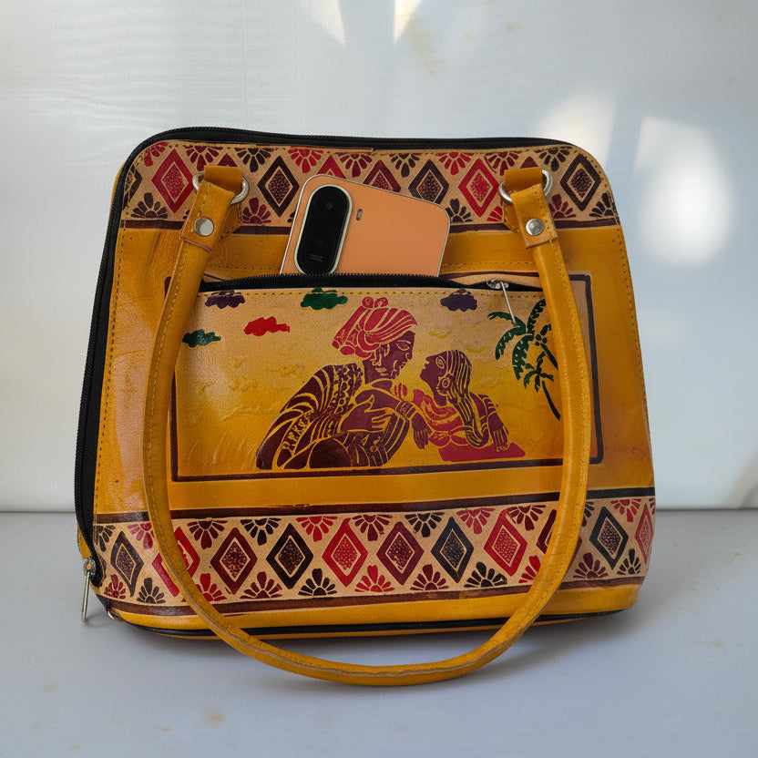 A mustard yellow handbag with a colorful hand-painted illustration of a couple on the front pocket, framed by geometric patterned borders. A peach-colored phone is tucked into the top pocket, and the bag is set against a light gray background with soft shadows.