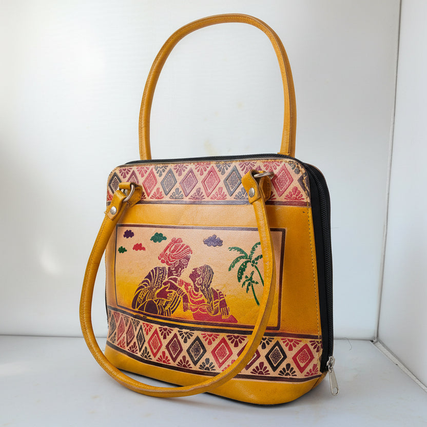 A mustard yellow handbag with a colorful hand-painted illustration of a couple under a palm tree on the front, framed by geometric patterned borders. The bag features a top handle, a shoulder strap, and silver-tone hardware, photographed against a white background.