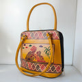 A mustard yellow handbag with a colorful hand-painted illustration of a couple under a palm tree on the front, framed by geometric patterned borders. The bag features a top handle, a shoulder strap, and silver-tone hardware, photographed against a white background.
