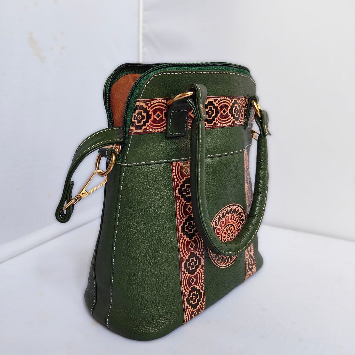 An olive green pebbled leather handbag with vertical orange and black patterned woven fabric panels, gold-tone hardware, and a top zipper, shown on a white surface against a white background.
