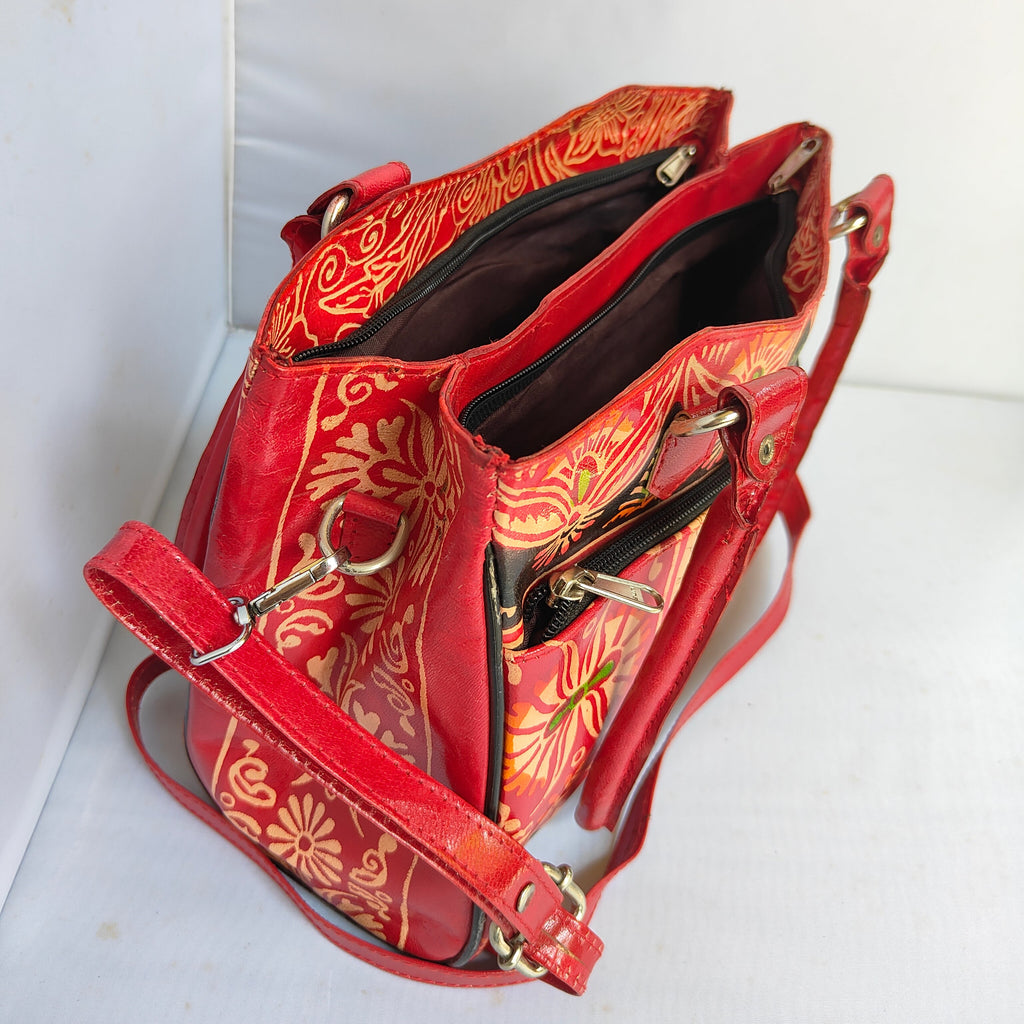 A red leather shoulder bag with a gold floral batik pattern, shown open to display its interior compartments and gold-tone hardware, set against a plain white background.