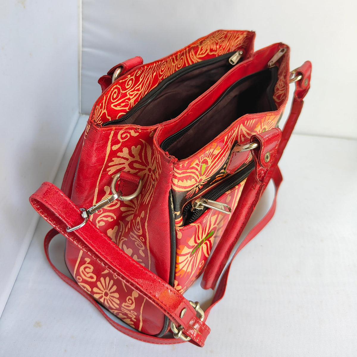 A red leather shoulder bag with a gold floral batik pattern, shown open to display its interior compartments and gold-tone hardware, set against a plain white background.