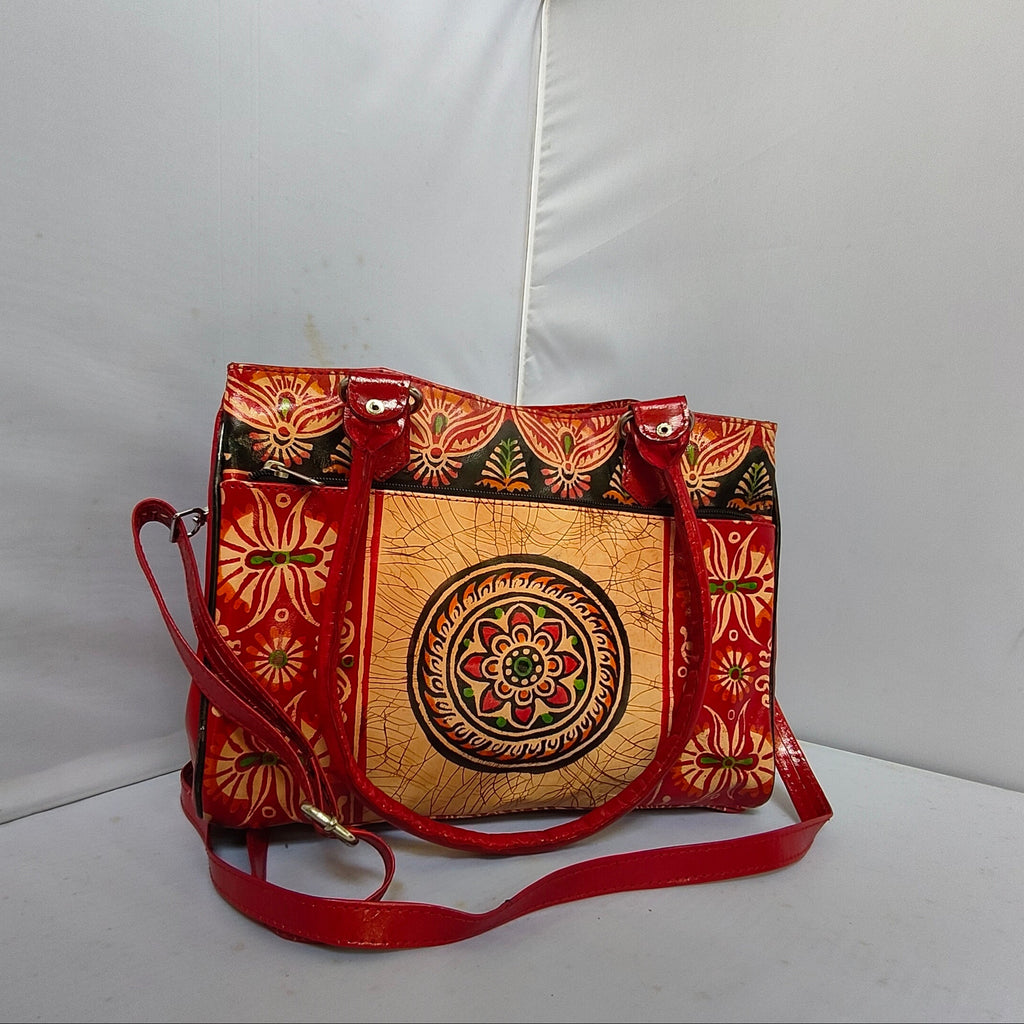 A red and gold patterned shoulder bag with a central mandala design on the front pocket, featuring a red leather strap and gold-tone hardware, set against a white studio backdrop.