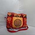 A red and gold patterned shoulder bag with a central mandala design on the front pocket, featuring a red leather strap and gold-tone hardware, set against a white studio backdrop.