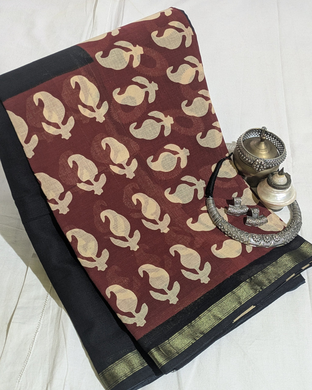 A folded dark red and black saree with beige motifs and a gold border, styled with traditional silver jewelry on a white fabric background.