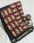 A folded dark red and black saree with beige motifs and a gold border, styled with traditional silver jewelry on a white fabric background.