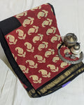 A folded red saree with beige floral motifs and a black border, displayed on a white fabric background alongside traditional silver oil lamps.