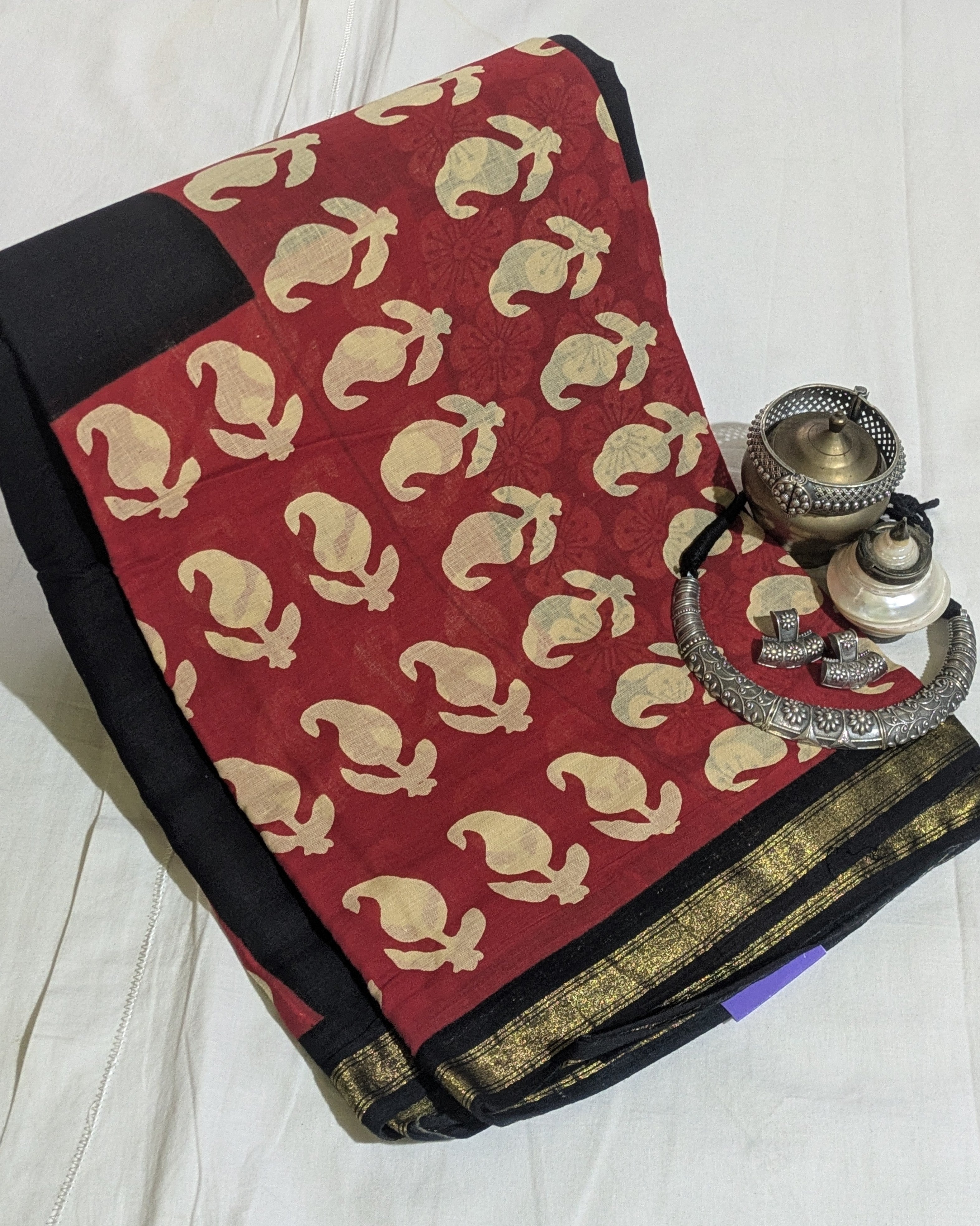 A folded red saree with beige floral motifs and a black border, displayed on a white fabric background alongside traditional silver oil lamps.