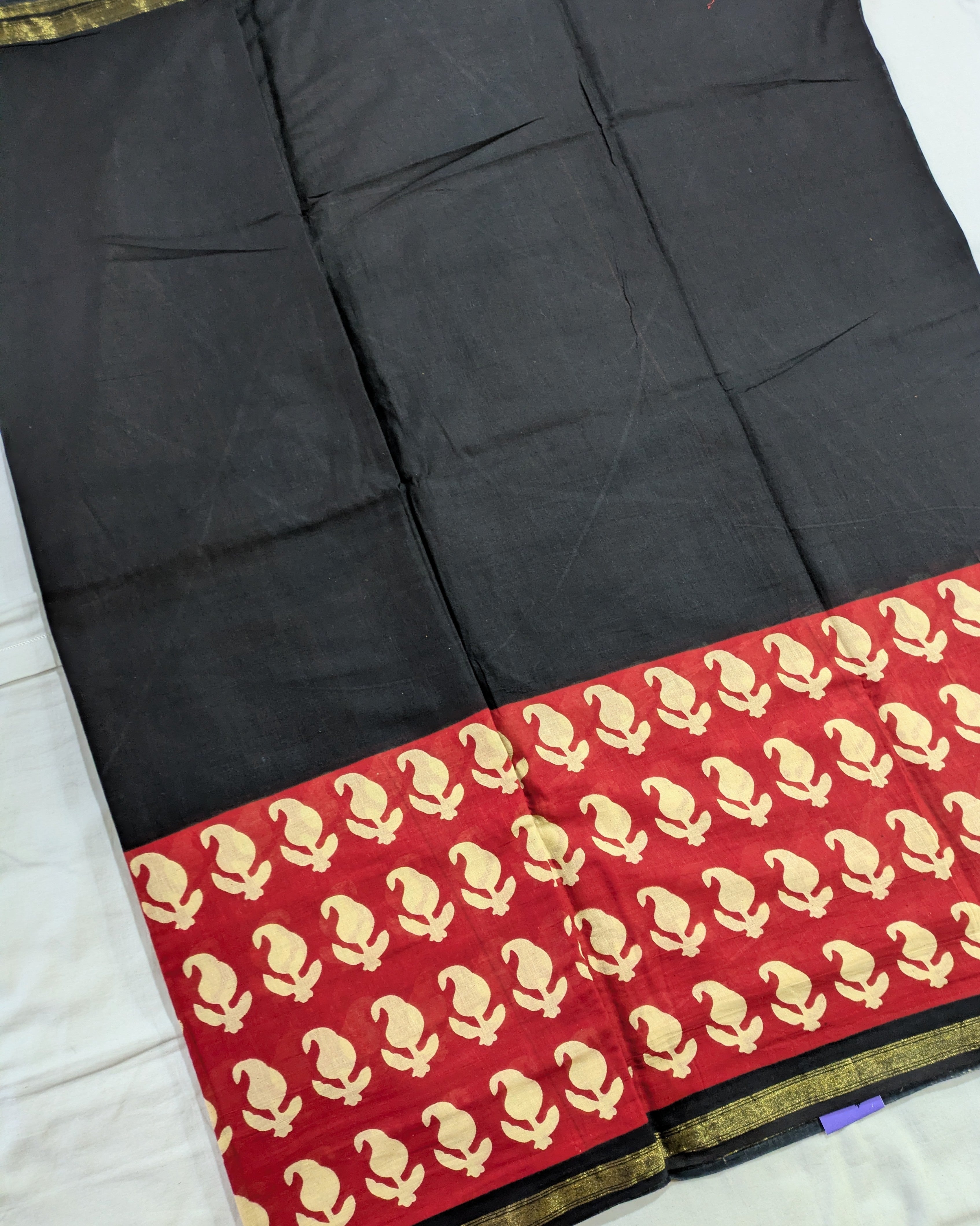 A black cotton saree or fabric panel with a wide red border featuring a repeating gold paisley pattern and a thin gold edge trim, laid flat on a white surface.