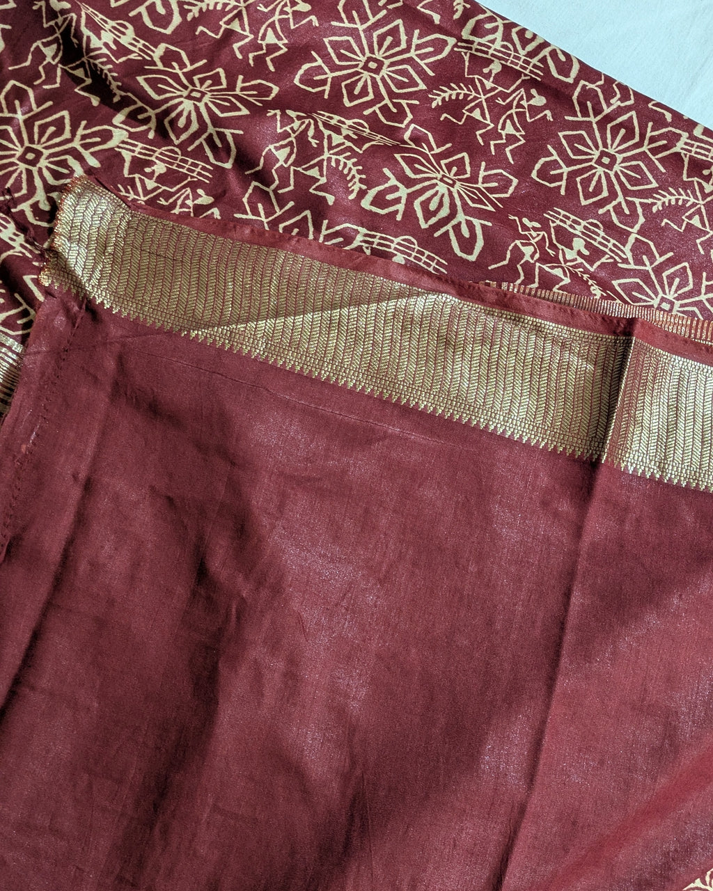 A close-up of a maroon fabric garment, likely a saree or dress, featuring a gold floral block print on the upper portion and a wide gold metallic border.