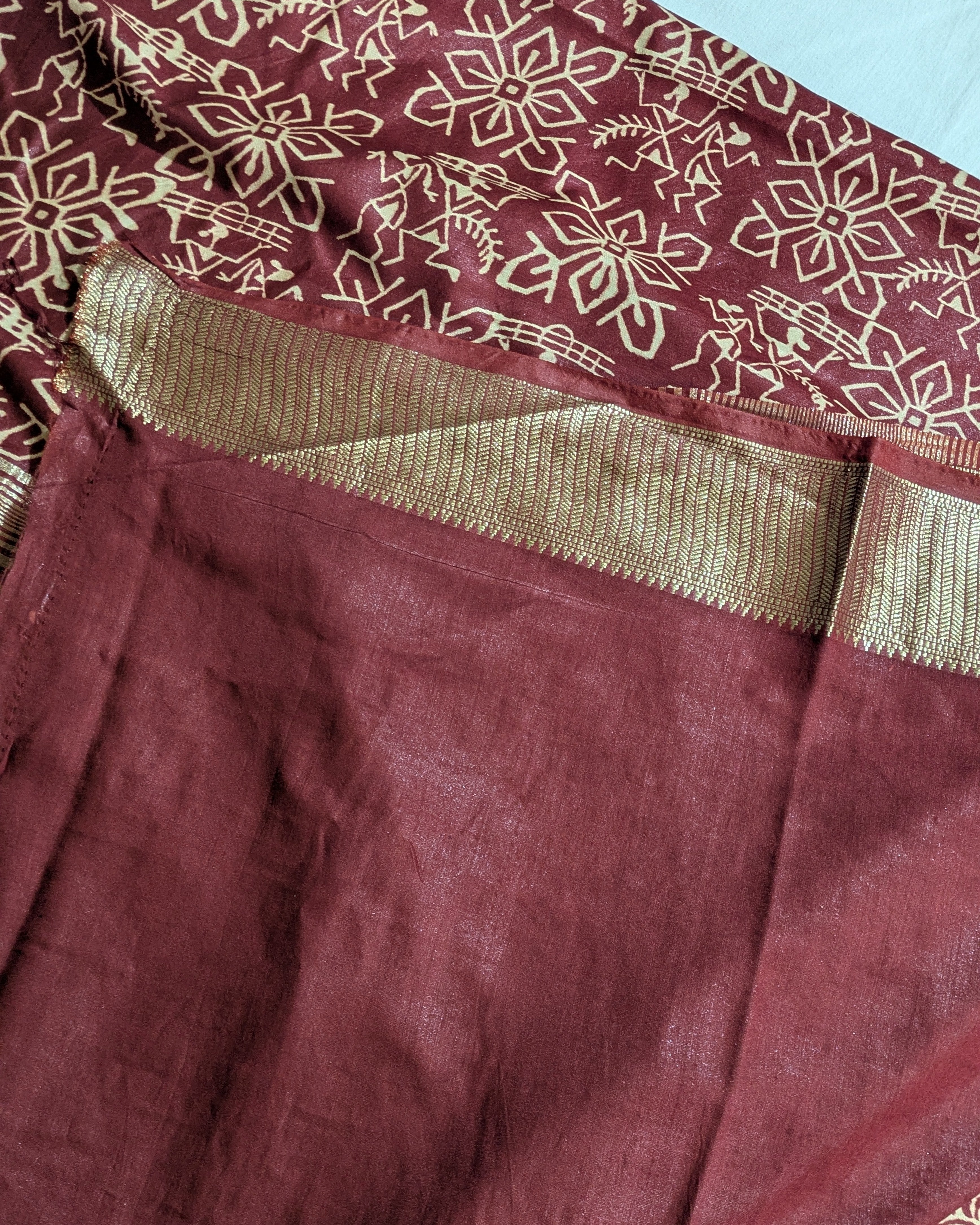 A close-up of a maroon fabric garment, likely a saree or dress, featuring a gold floral block print on the upper portion and a wide gold metallic border.
