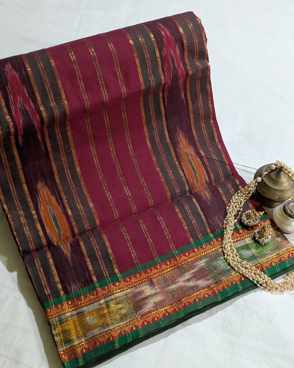 A folded traditional Indian saree with a deep magenta-pink body, featuring vertical stripes of gold, brown, and orange, and a wide green and gold border. The saree is styled on a white fabric background with decorative brass and pearl accessories.