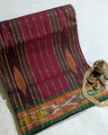A folded traditional Indian saree with a deep magenta-pink body, featuring vertical stripes of gold, brown, and orange, and a wide green and gold border. The saree is styled on a white fabric background with decorative brass and pearl accessories.
