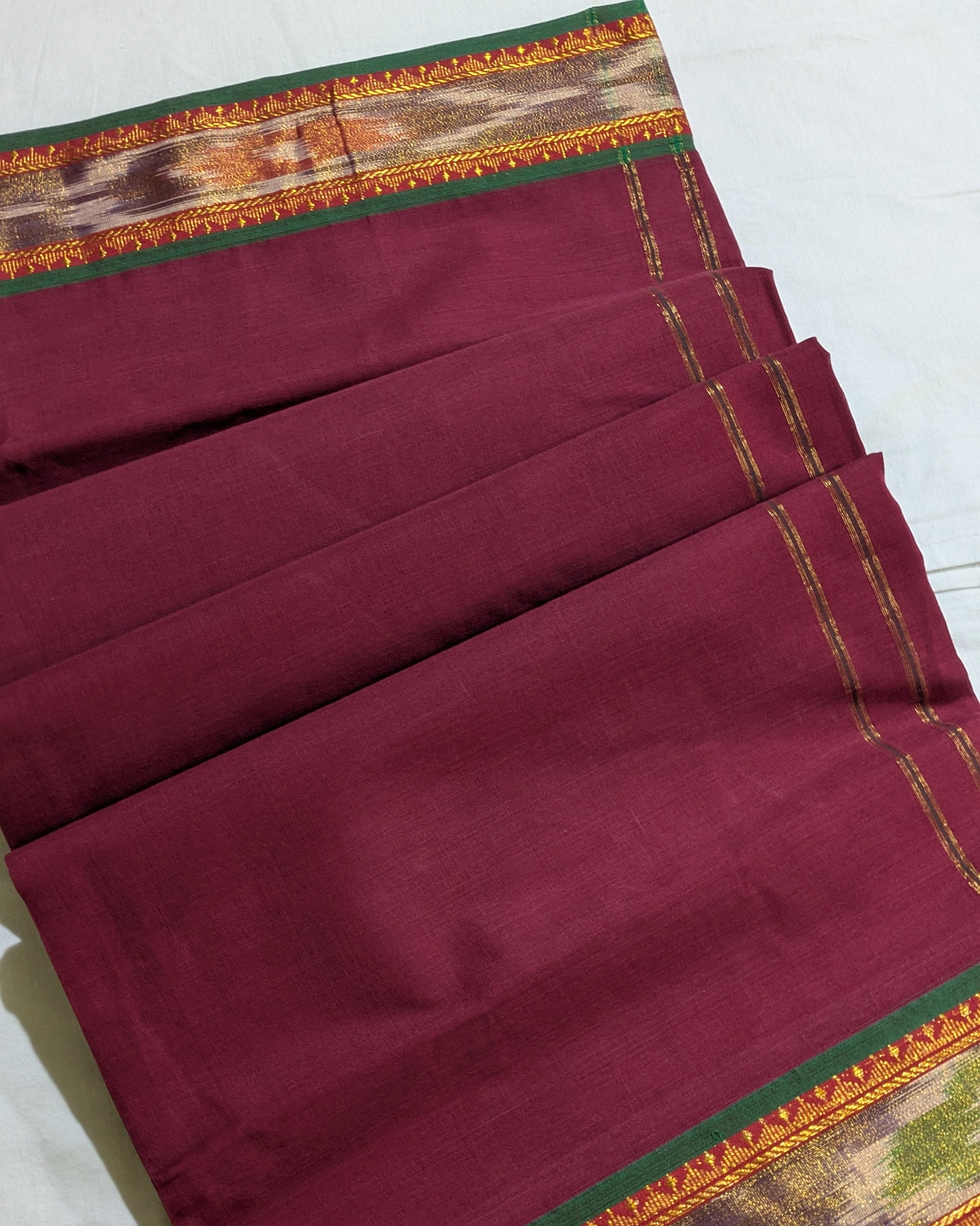 A folded maroon silk saree with a detailed multicolor border featuring green, gold, and brown patterns, laid out on a light-colored surface.