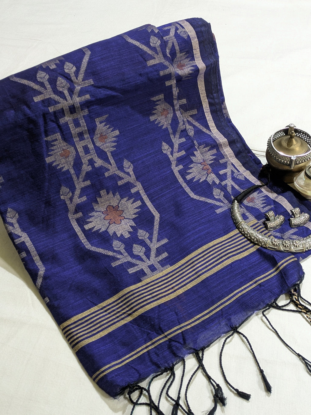 A folded royal blue silk saree or scarf with intricate white floral motifs, gold stripes, and black tassels, styled on a white fabric background next to traditional brass and silver accessories.
