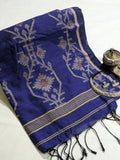 A folded royal blue silk saree or scarf with intricate white floral motifs, gold stripes, and black tassels, styled on a white fabric background next to traditional brass and silver accessories.