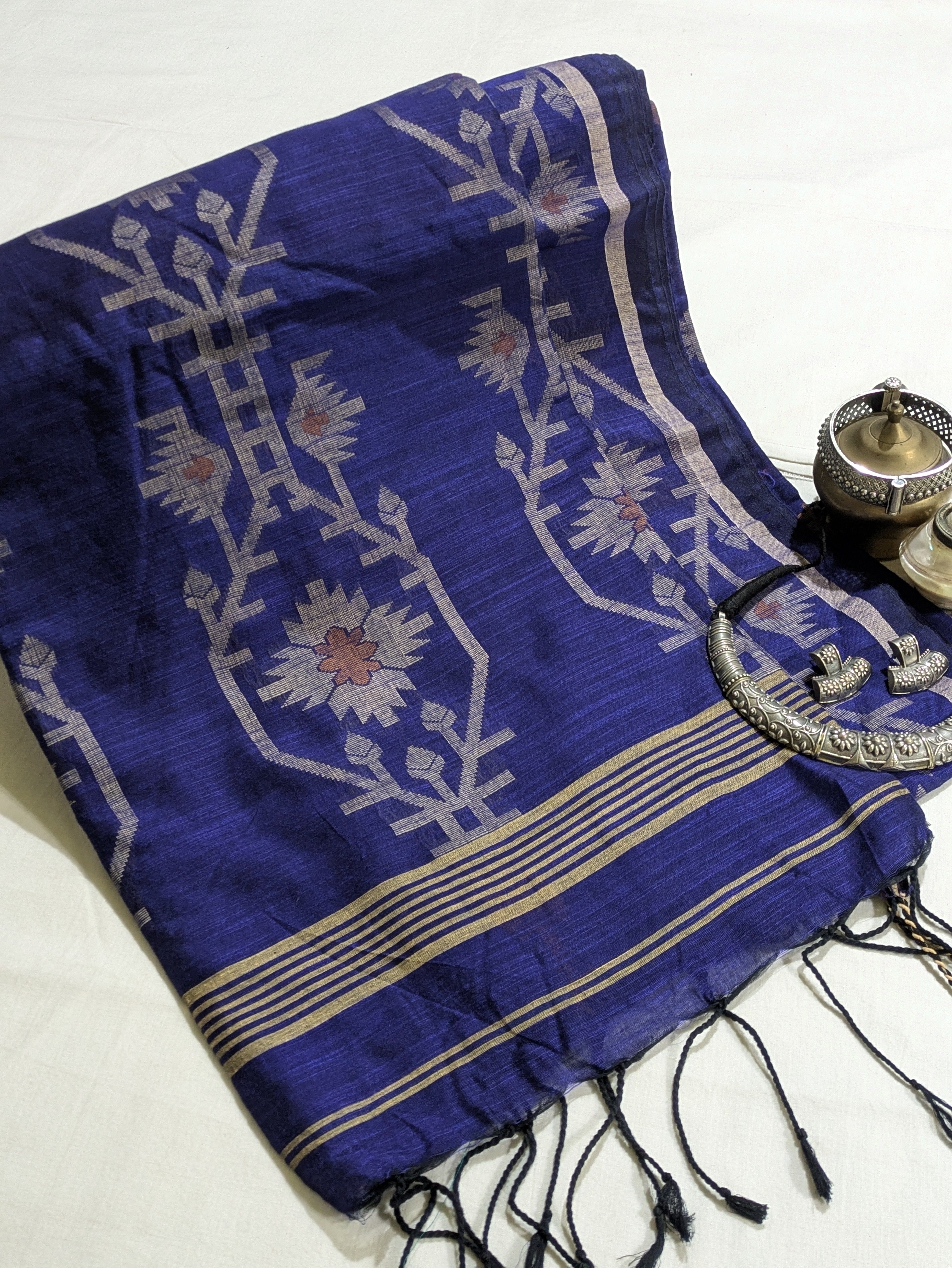 A folded royal blue silk saree or scarf with intricate white floral motifs, gold stripes, and black tassels, styled on a white fabric background next to traditional brass and silver accessories.