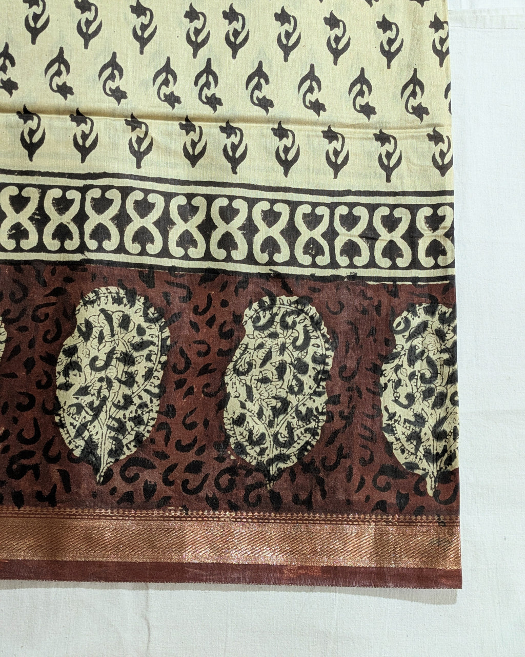 A close-up of a traditional Indian fabric, likely silk or satin, featuring a cream-colored upper section with black block prints, a wide decorative border, and a rich brown lower section with three large paisley motifs and a metallic gold border.