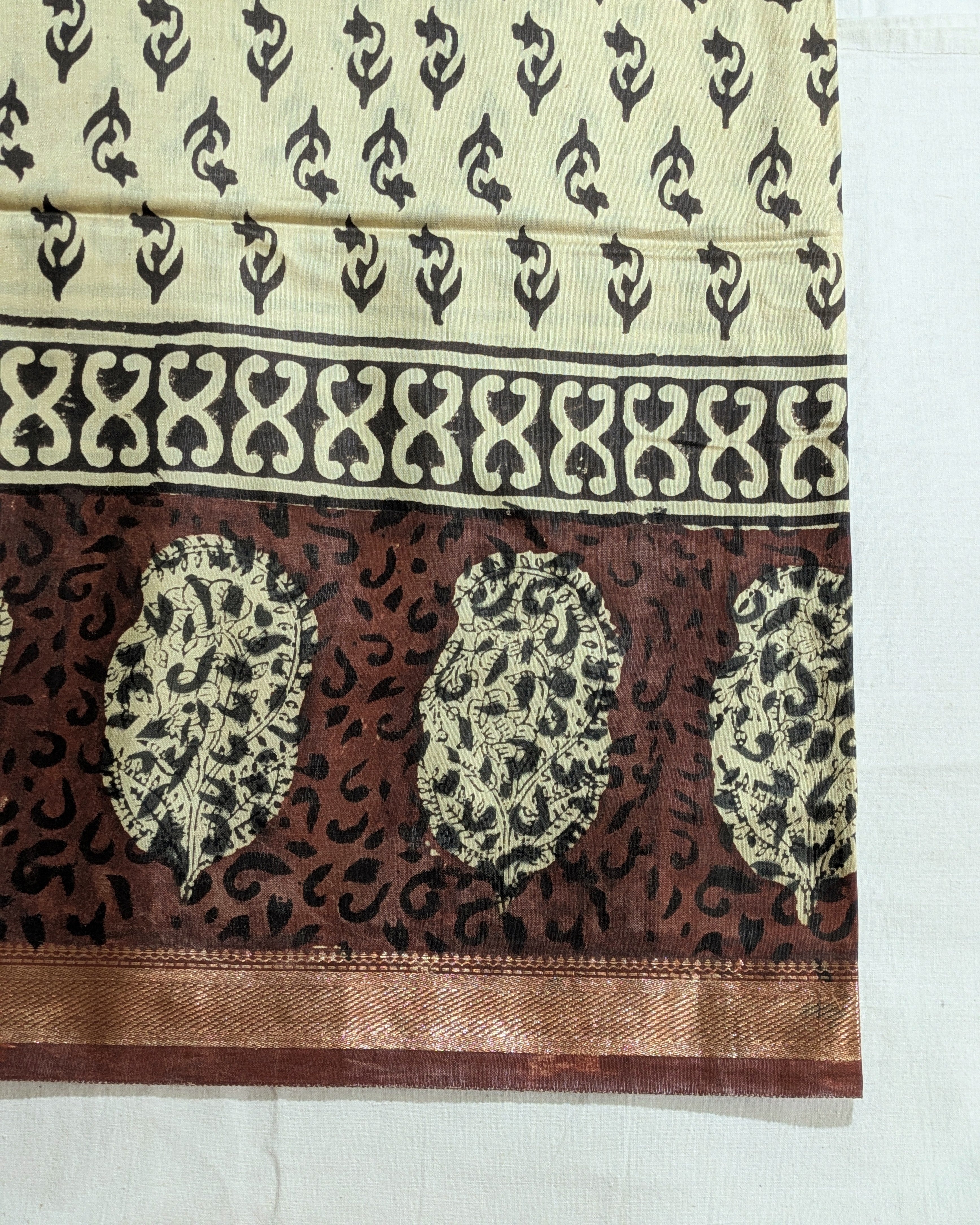 A close-up of a traditional Indian fabric, likely silk or satin, featuring a cream-colored upper section with black block prints, a wide decorative border, and a rich brown lower section with three large paisley motifs and a metallic gold border.