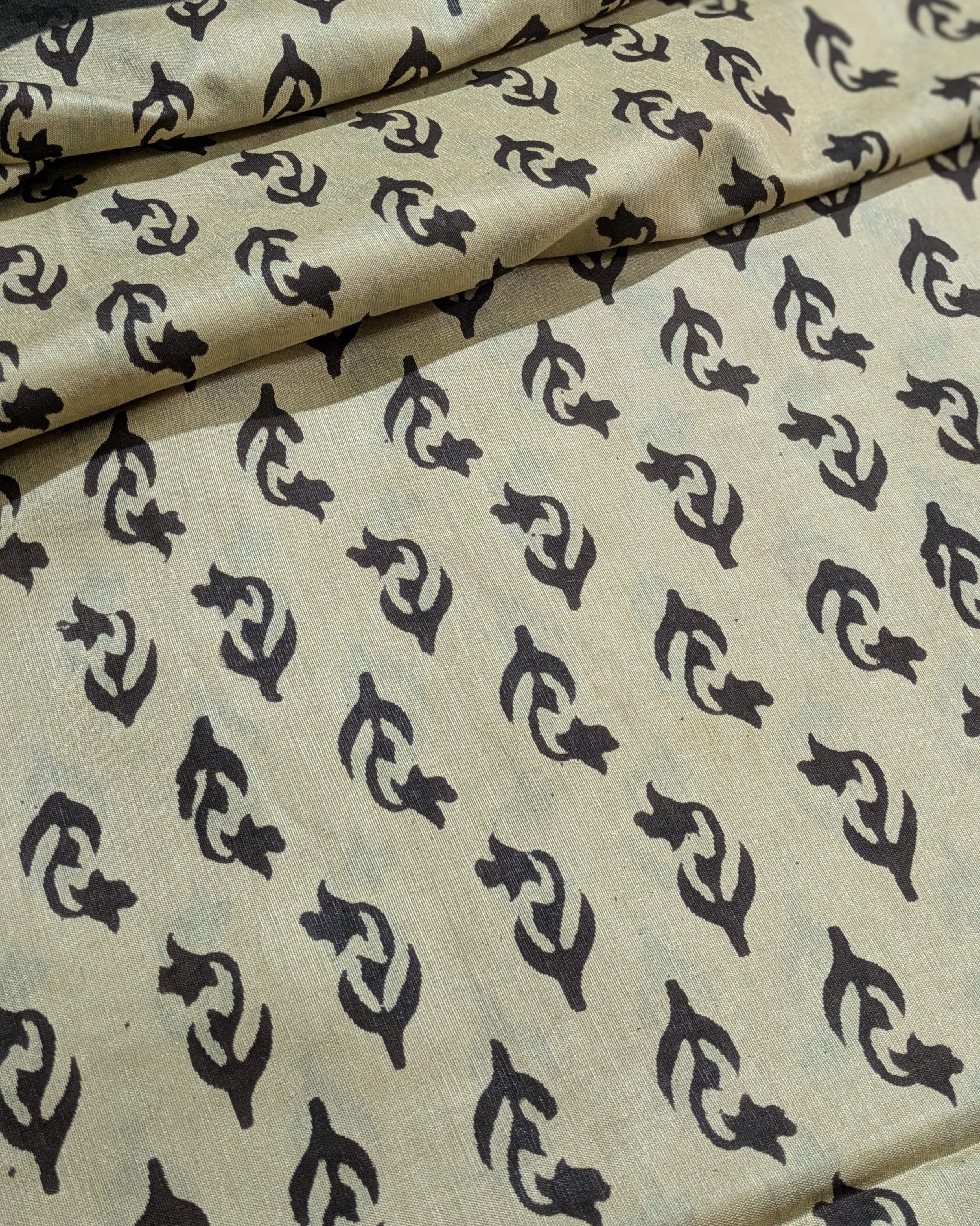 Beige fabric with a repeating black block print pattern of stylized fish or whale-like motifs, shown in soft folds.