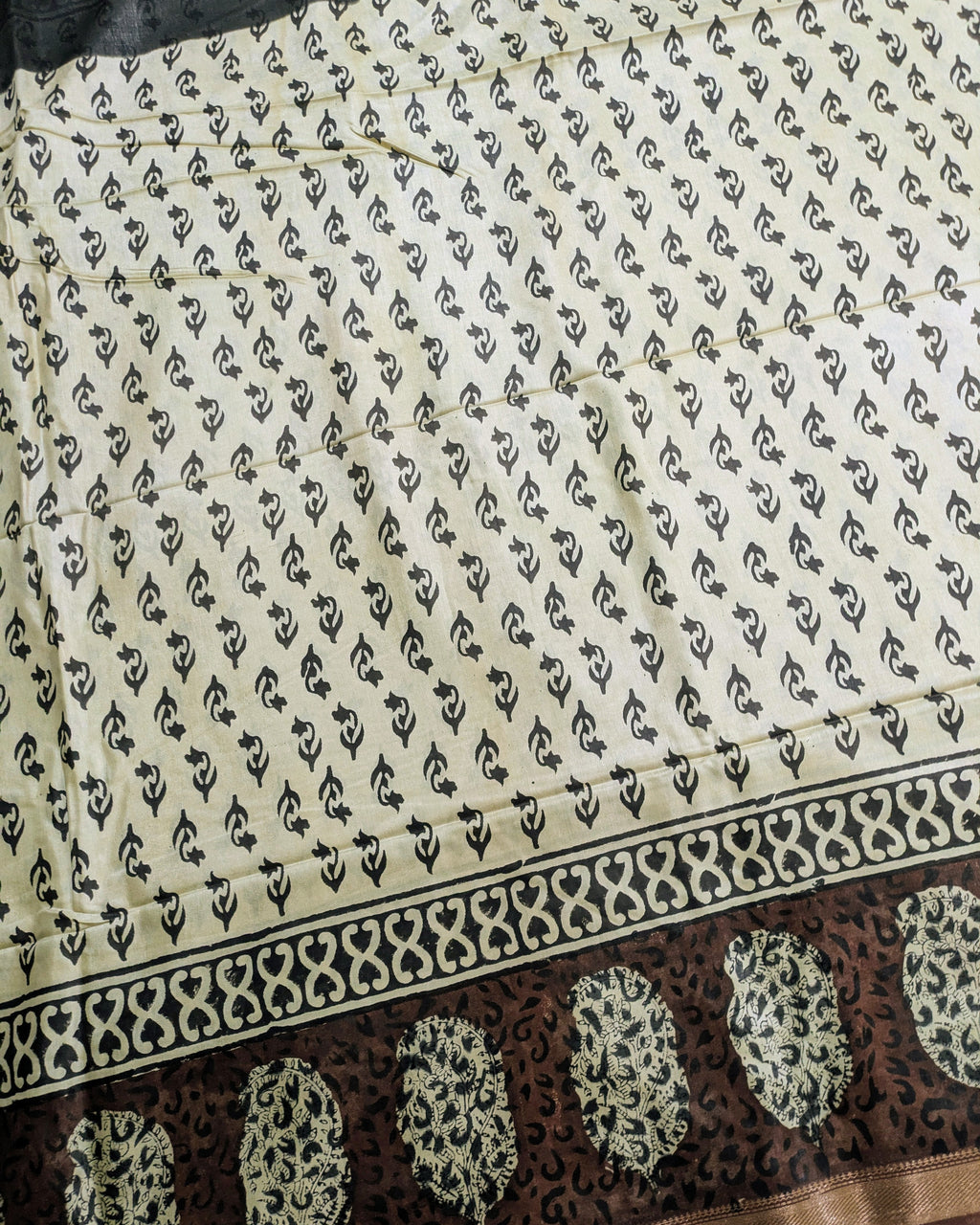 A close-up of a printed fabric, likely a saree, featuring a cream-colored body with a repeating black block print pattern and a wide dark brown border with cream floral motifs.