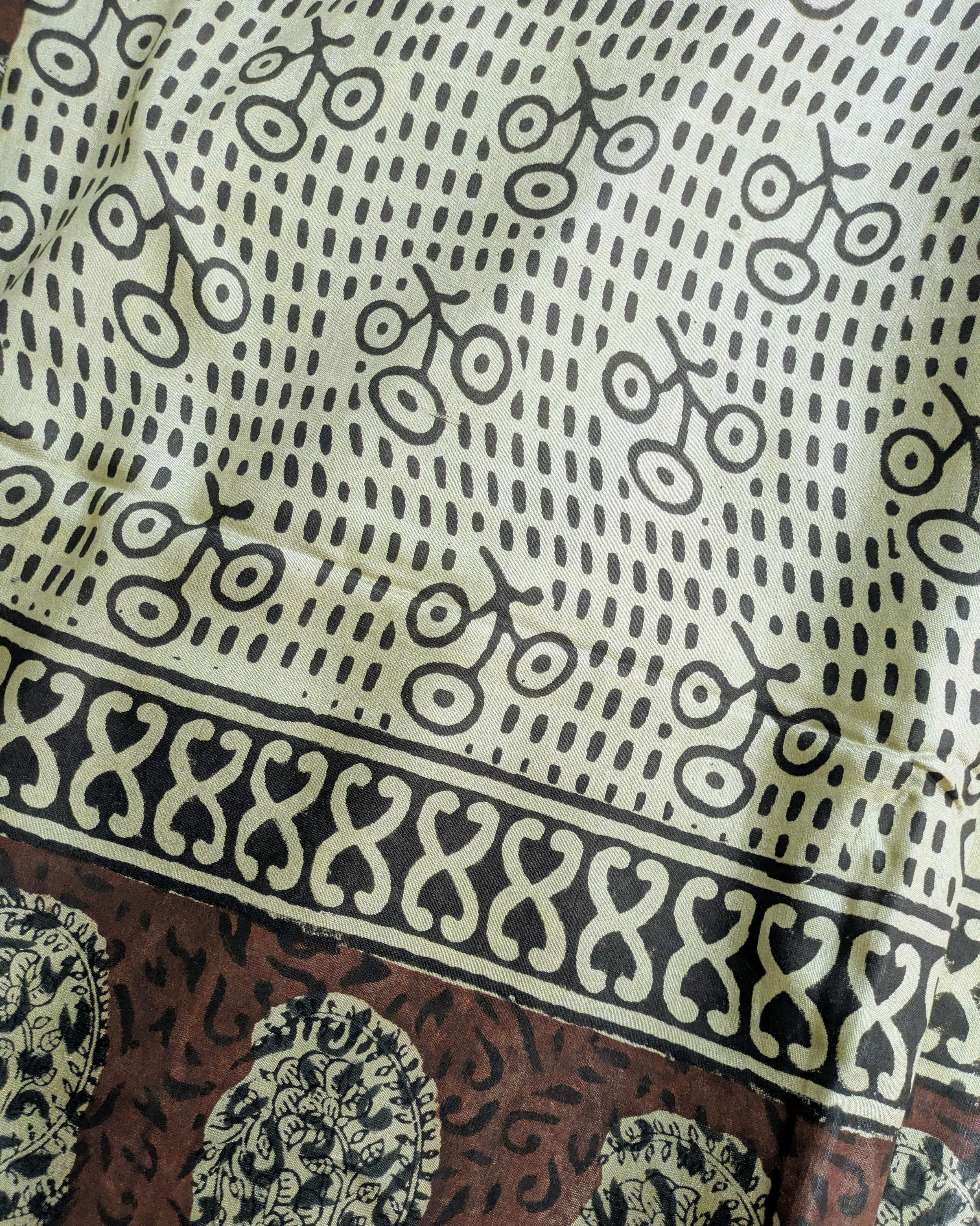 A close-up of a printed textile showing a cream and black patterned upper section with repeated bicycle-like motifs, a black decorative border, and a brown lower section with ornate paisley-style prints.