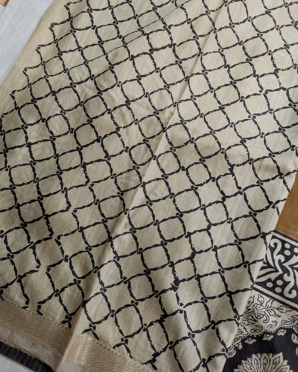 A close-up of a beige fabric with a black geometric lattice pattern, showing a woven border and a small portion of a patterned textile in the bottom right corner.
