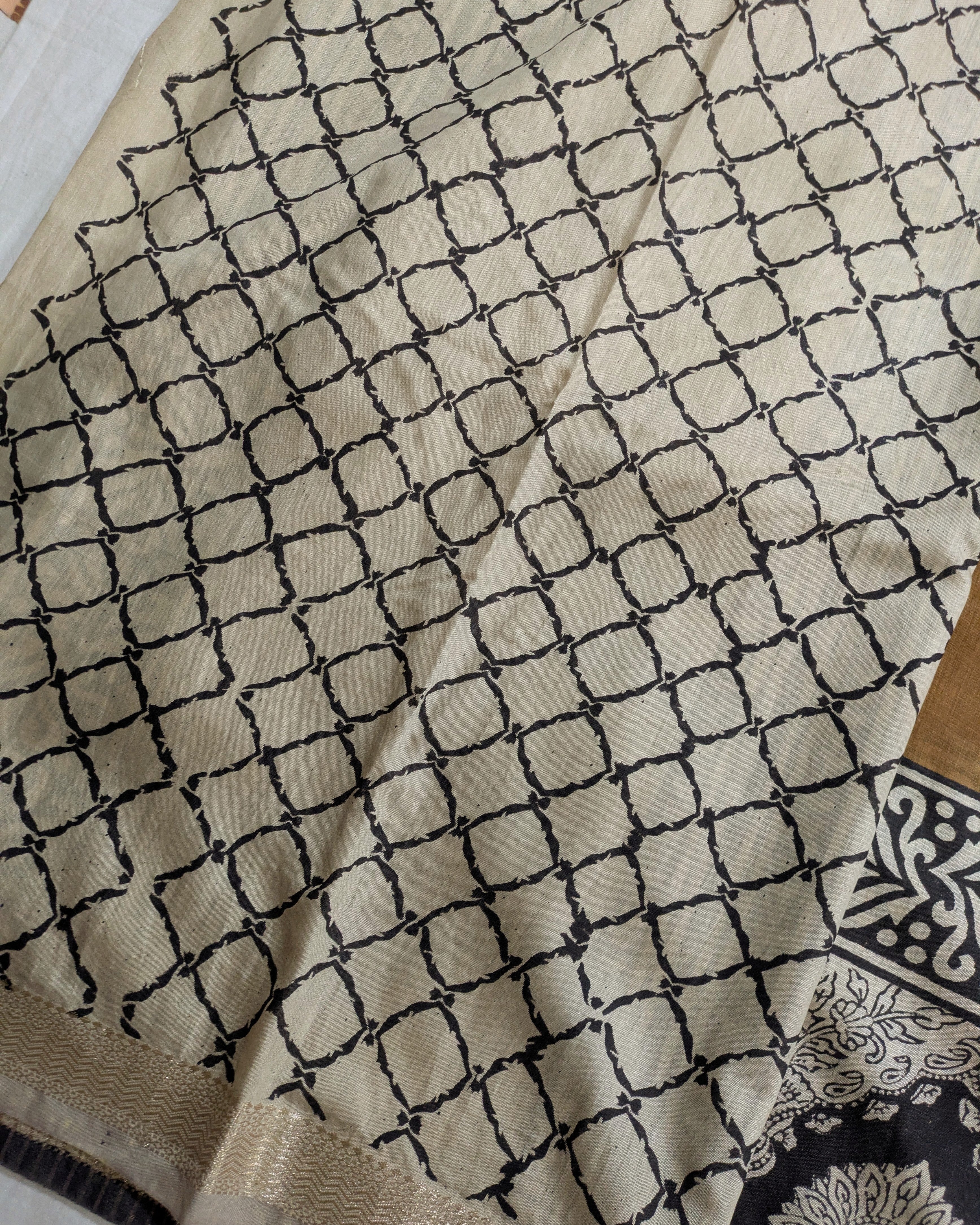 A close-up of a beige fabric with a black geometric lattice pattern, showing a woven border and a small portion of a patterned textile in the bottom right corner.