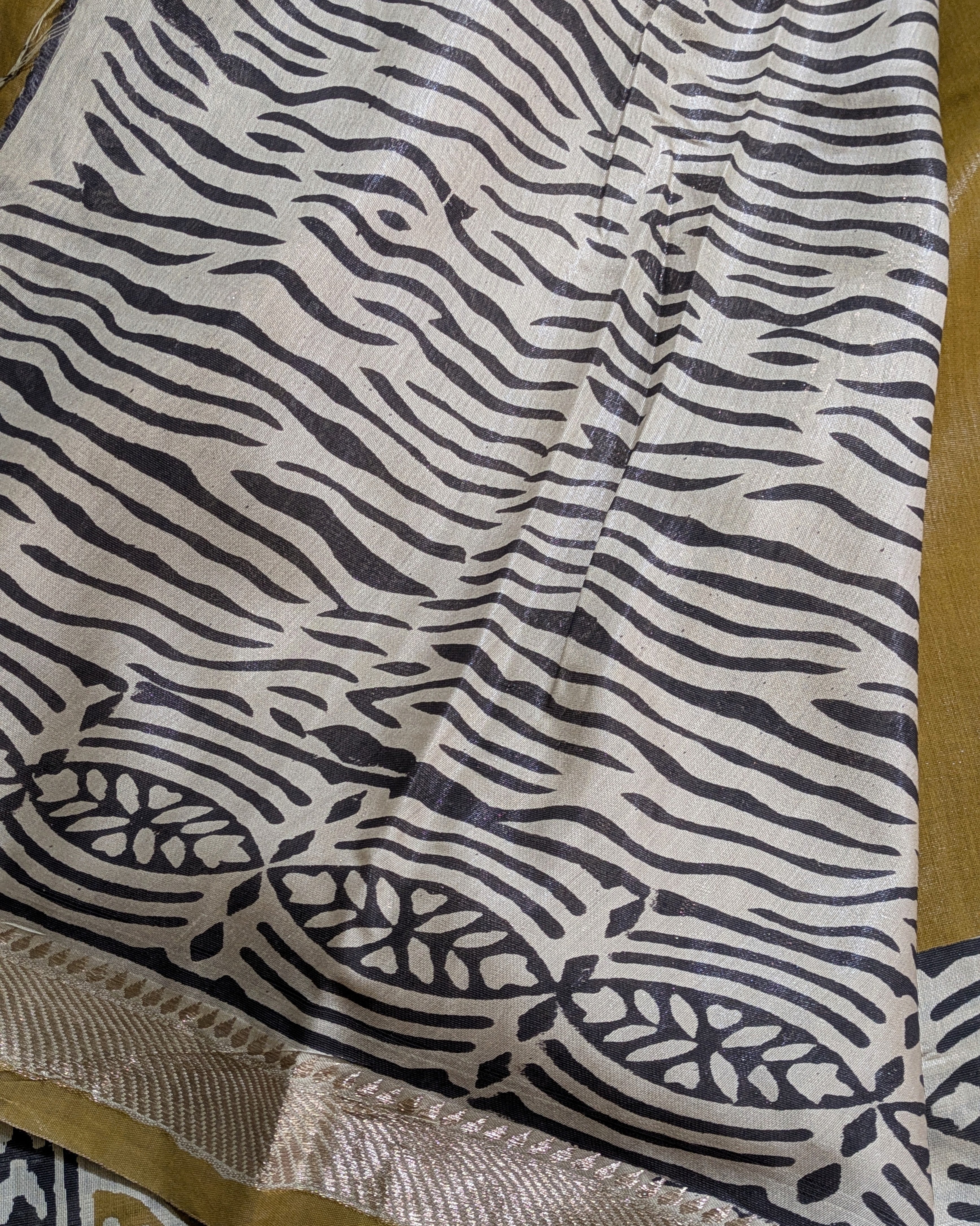 A close-up of a fabric textile with a black zebra-like wavy stripe pattern on an off-white base, plus a decorative border with floral motifs and a metallic-looking beige edge.