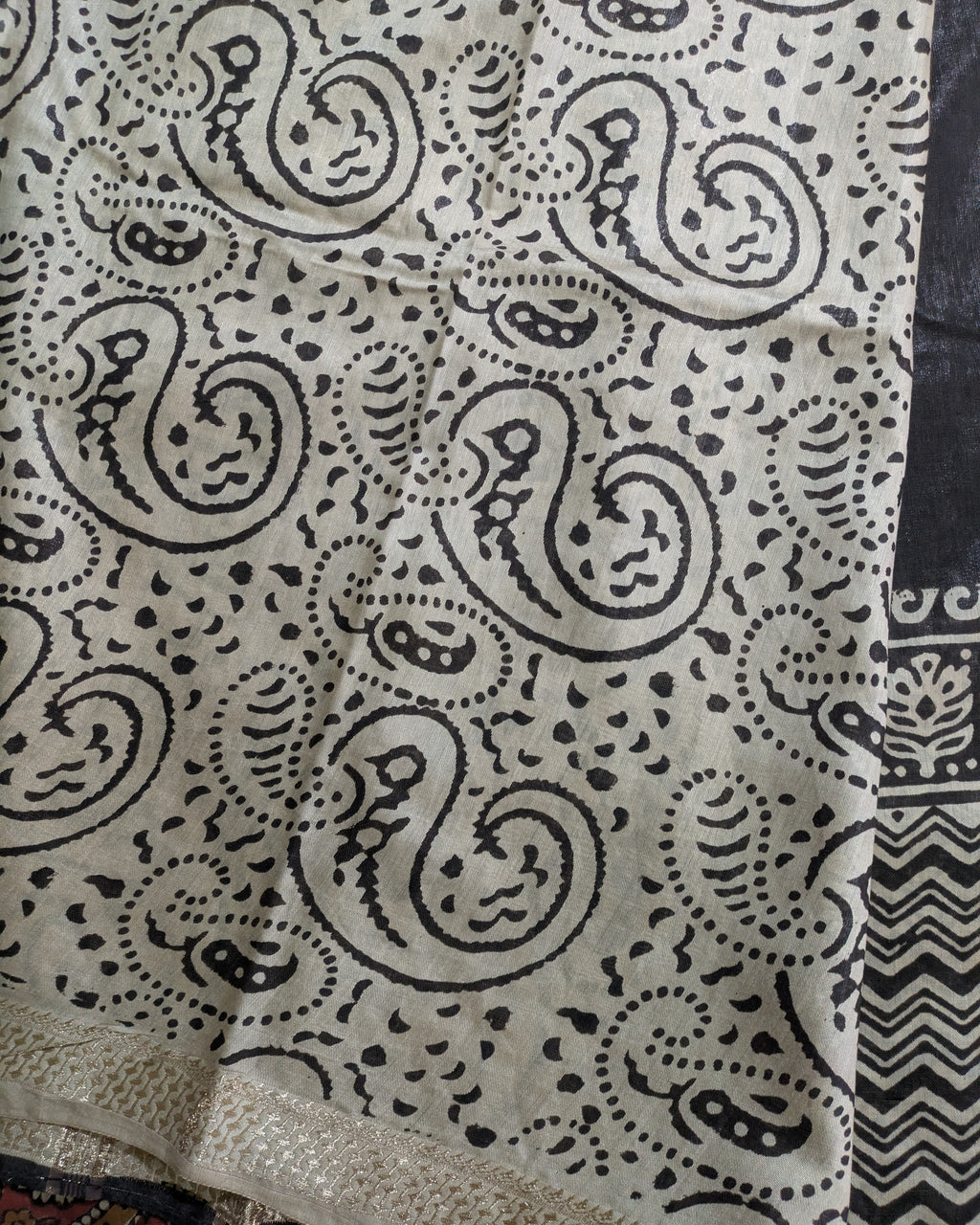 A close-up of a fabric with a black paisley and dot pattern on a light beige background, showing a textured woven border at the bottom and a patterned section on the right.