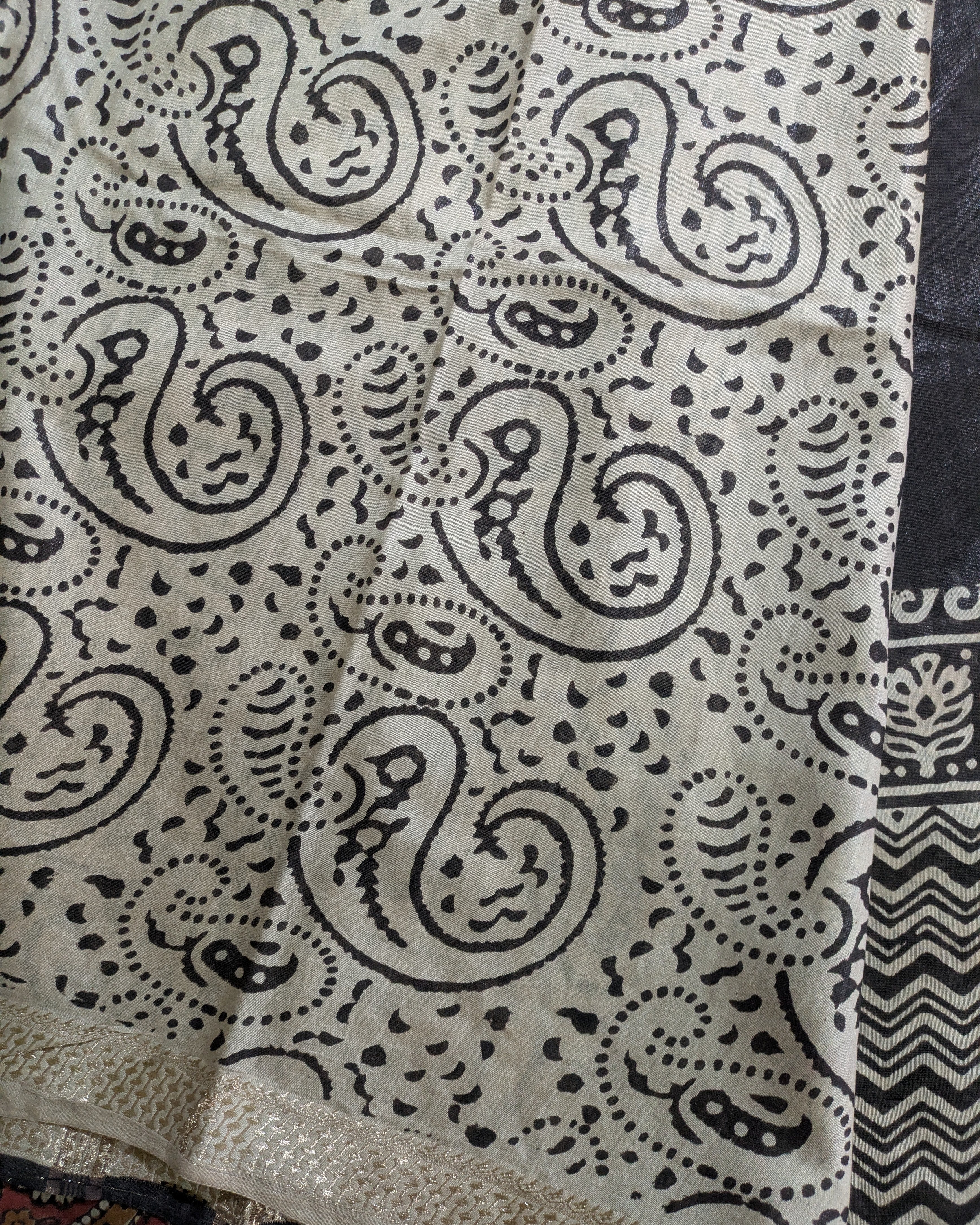 A close-up of a fabric with a black paisley and dot pattern on a light beige background, showing a textured woven border at the bottom and a patterned section on the right.