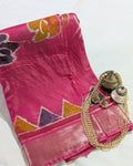 A folded pink silk saree with a metallic zari border, featuring orange and purple floral motifs, styled with traditional jewelry on a white background.
