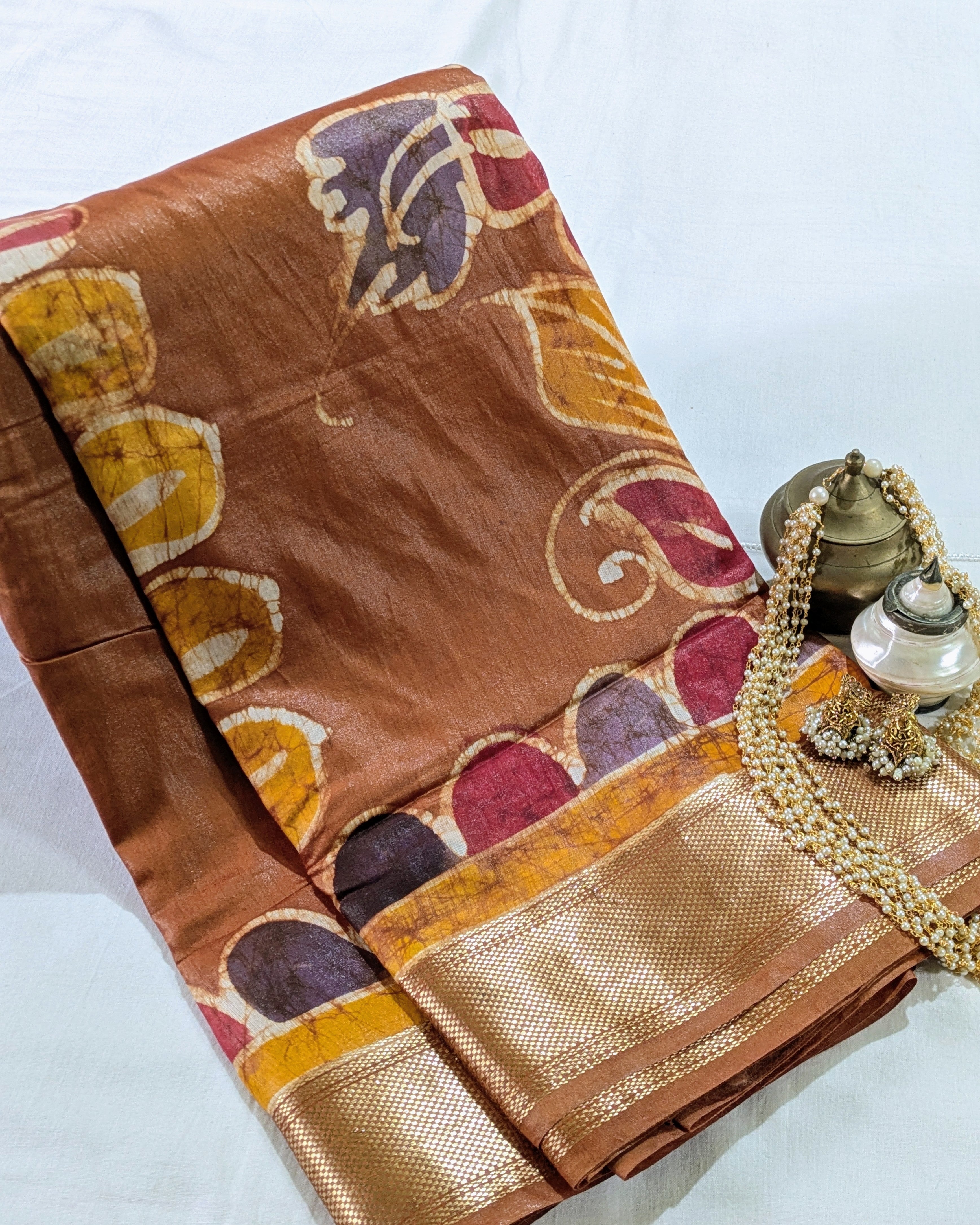 A folded brown silk saree with a wide gold zari border and colorful hand-dyed floral motifs in yellow, red, and purple, styled on a white fabric background with traditional decorative props.