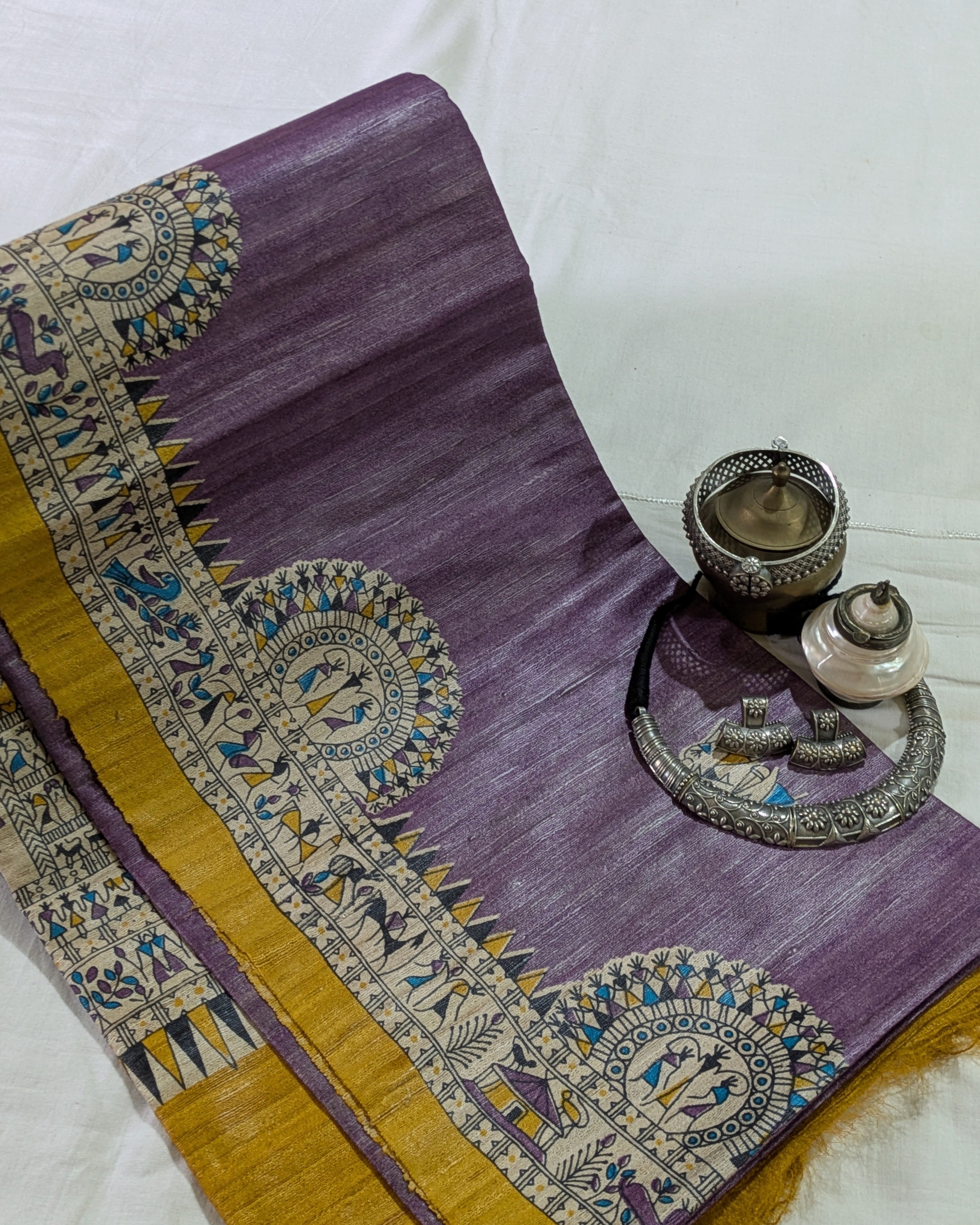 A purple silk saree with a wide gold border featuring intricate blue, yellow, and white traditional motifs, styled with silver jewelry on a white fabric background.