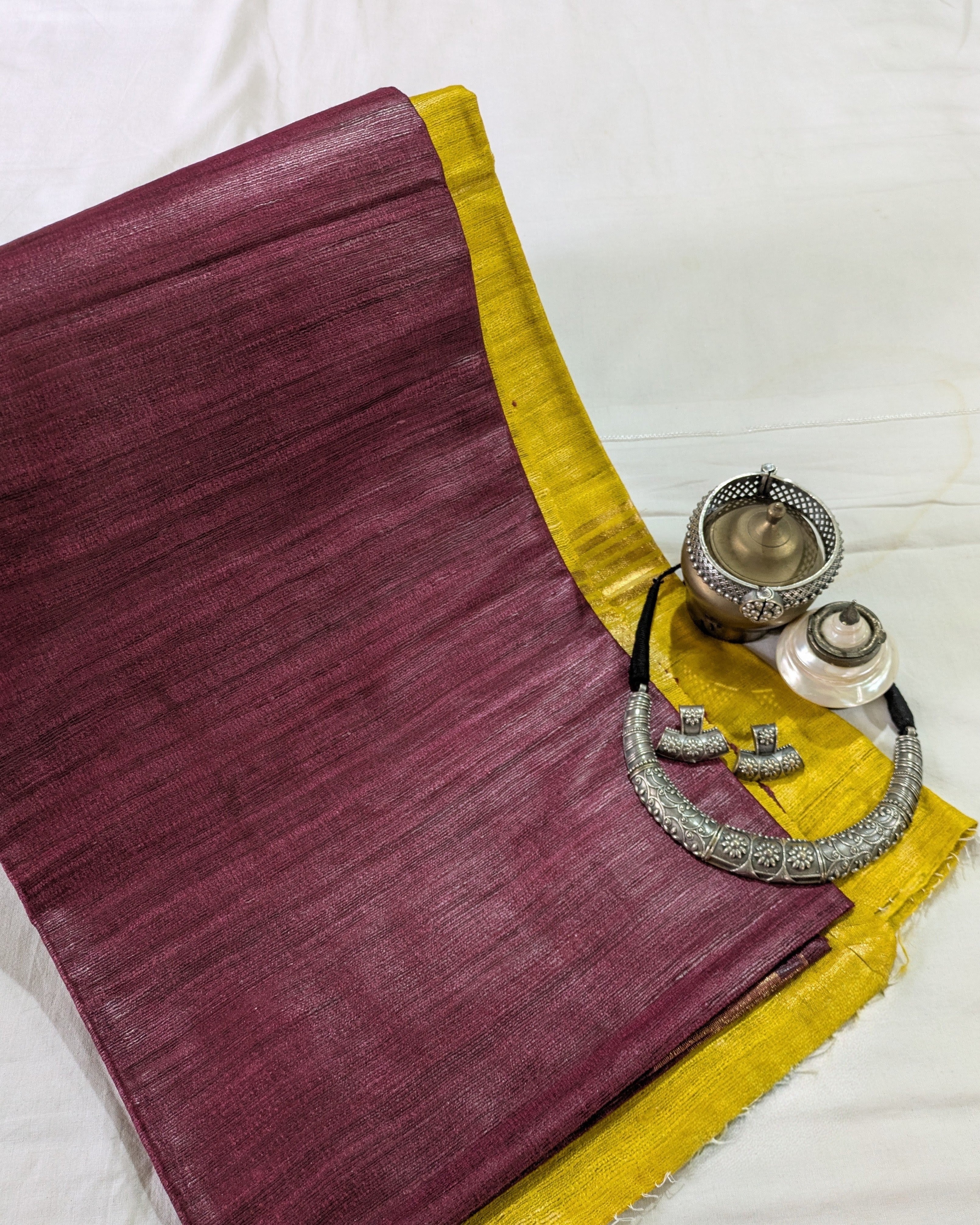 A folded maroon silk saree with a yellow border, styled with traditional silver jewelry on a white fabric background.