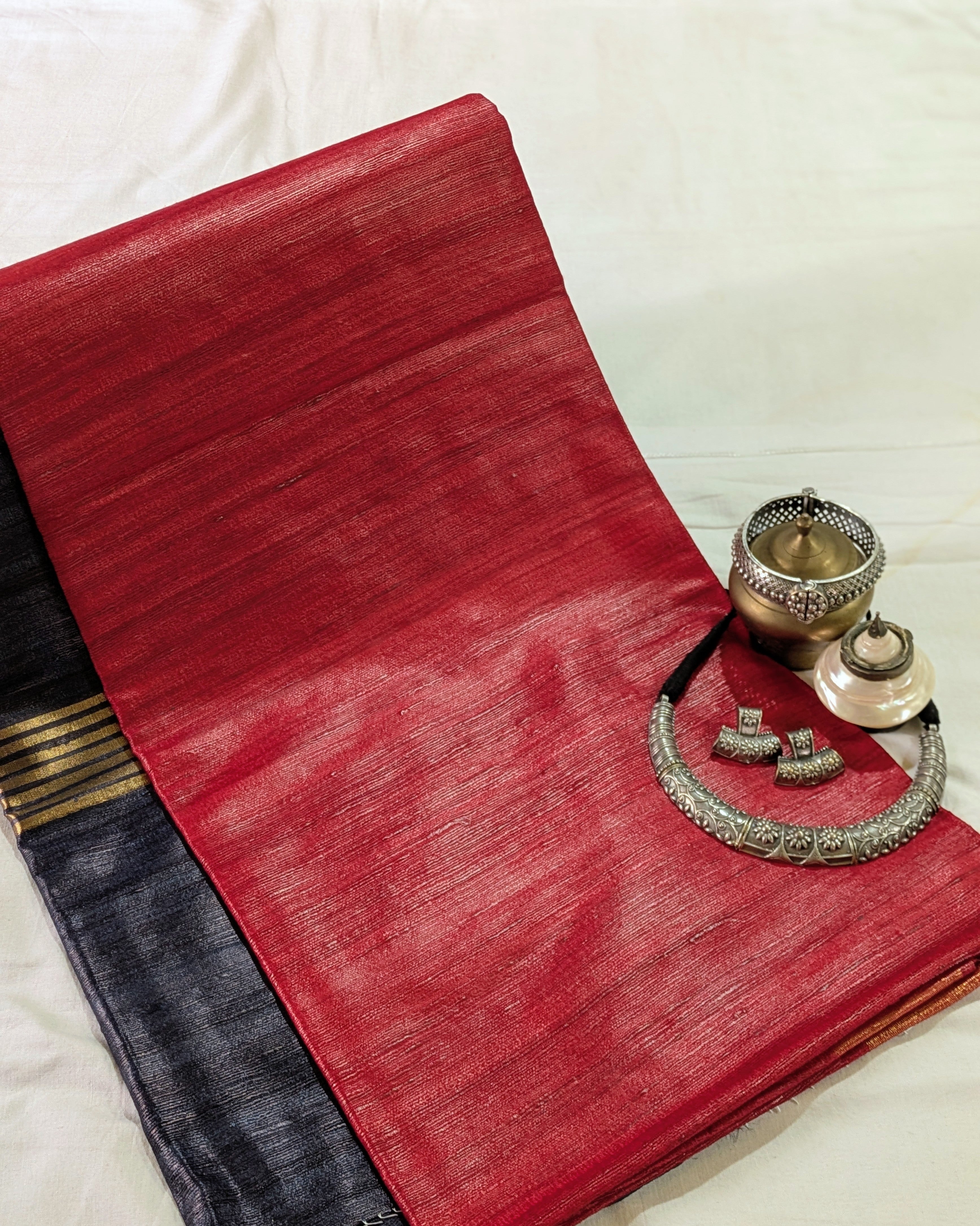 A folded red silk saree with a dark navy border and gold stripes, styled with traditional Indian jewelry on a white fabric background.