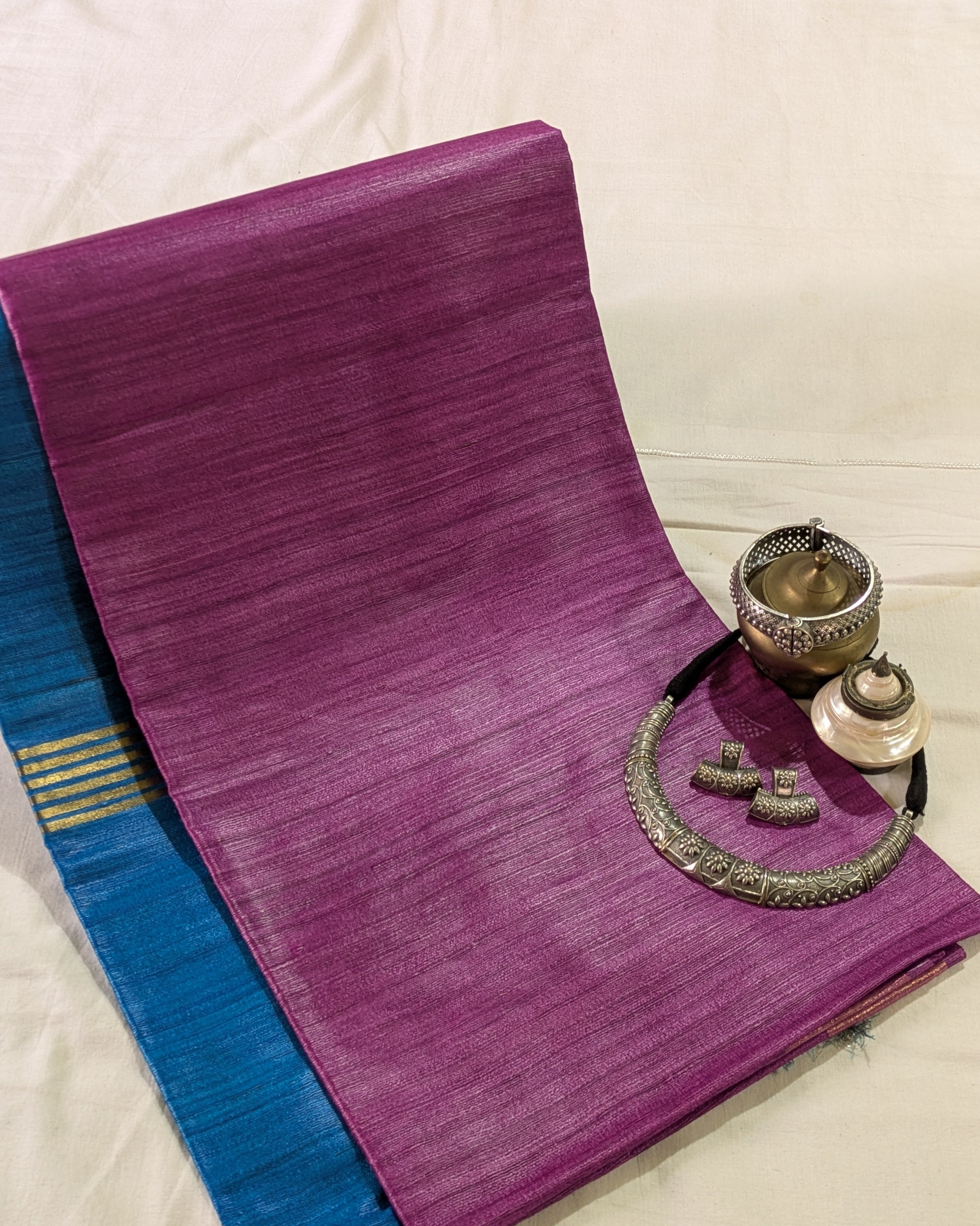 A folded purple silk saree with a blue border and gold stripes, styled with traditional silver jewelry on a white fabric background.