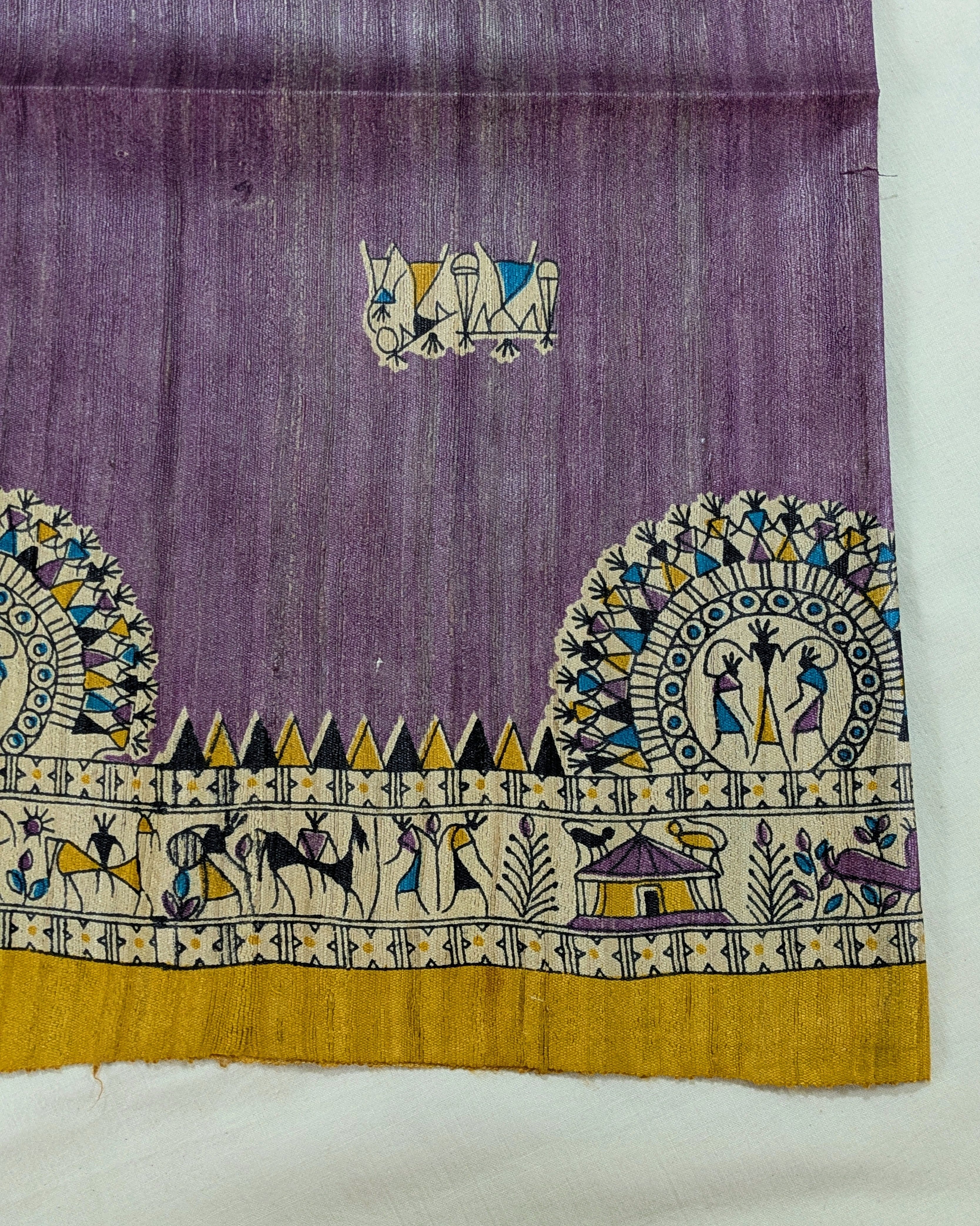 A close-up of a purple textile with a wide decorative border featuring cream, yellow, blue, and black folk-art motifs, including animals, geometric patterns, and small house illustrations.