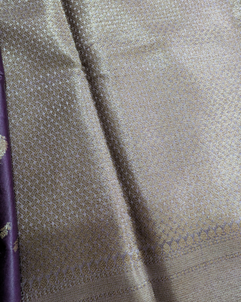 A close-up of a woven fabric with a shimmering metallic silver body and intricate gold zari embroidery, showing a subtle purple border on the left edge.