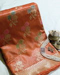 An orange-pink Banarasi silk saree with woven floral motifs in gold, pink, and green, styled with pearl jewelry on brass vessels against a white fabric background.