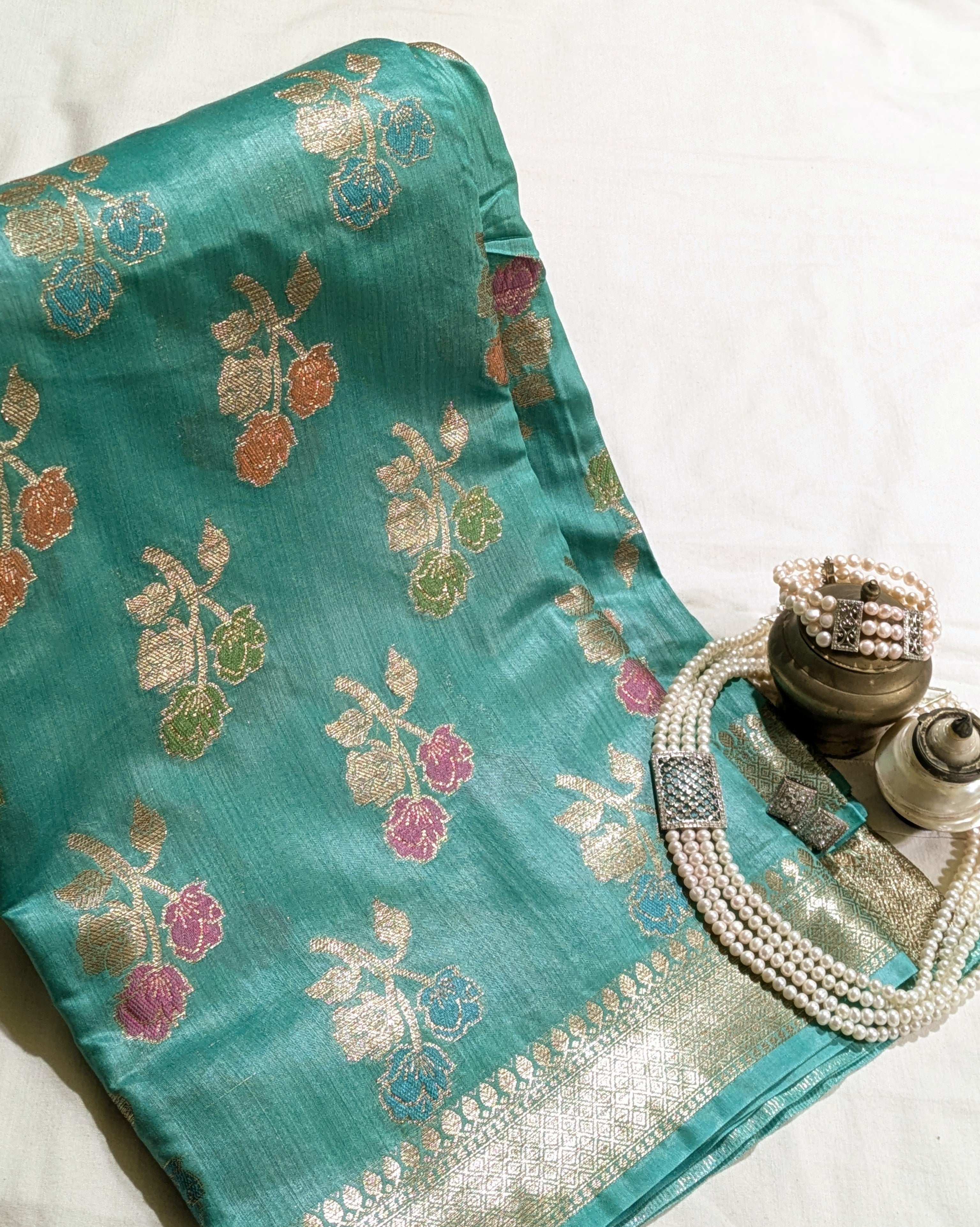 A teal green silk saree with intricate floral motifs in gold, pink, and green, styled with a pearl necklace and brass vessels on a white fabric background.